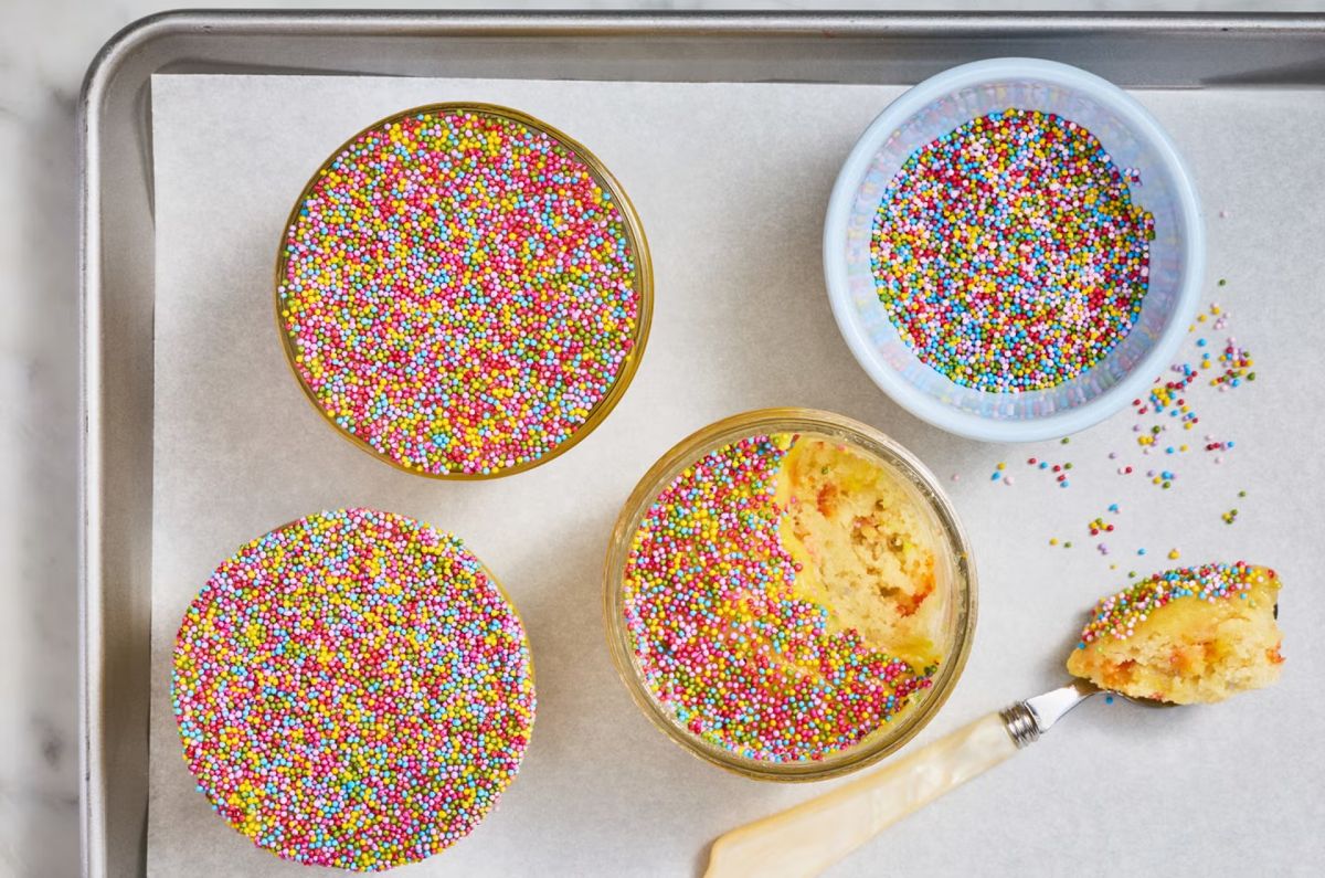 Individual cakes covered edge to edge in rainbow nonpareils on a baking tray, one opened to show the soft crumb