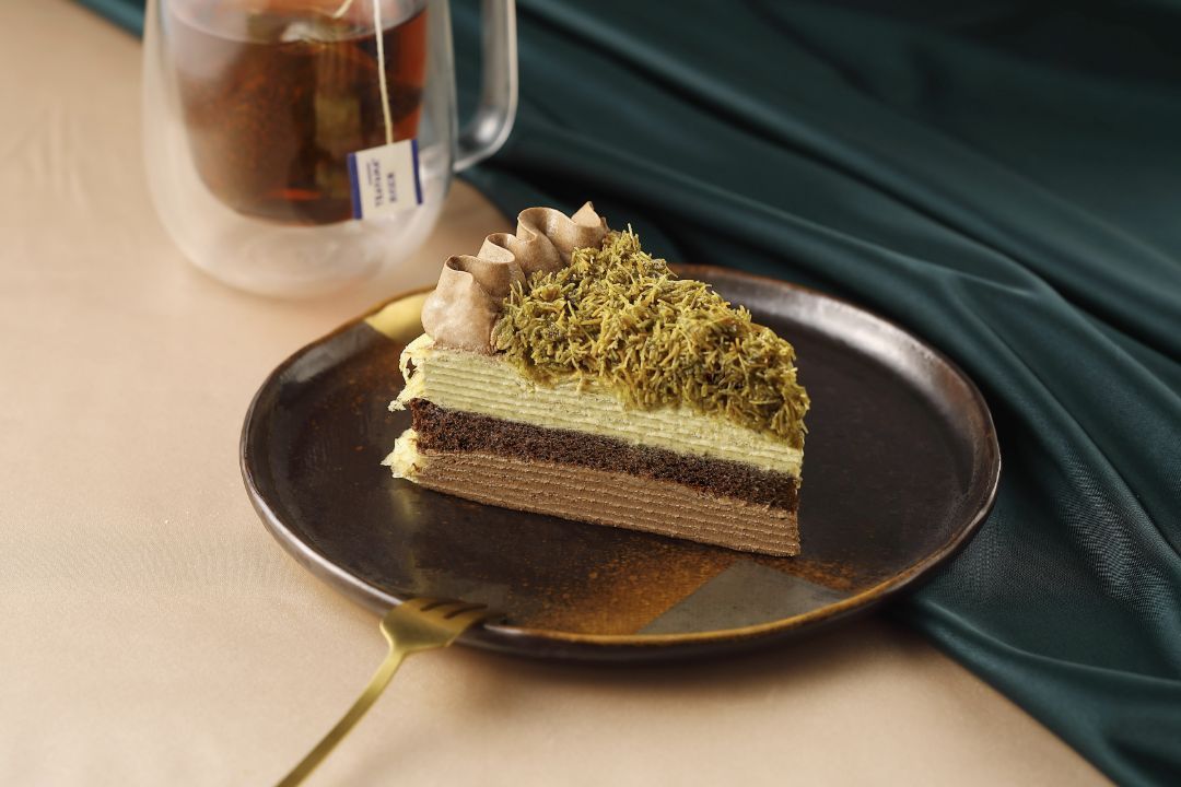 A slice of layered chocolate mille-crêpe topped with green pistachio strands on a dark plate