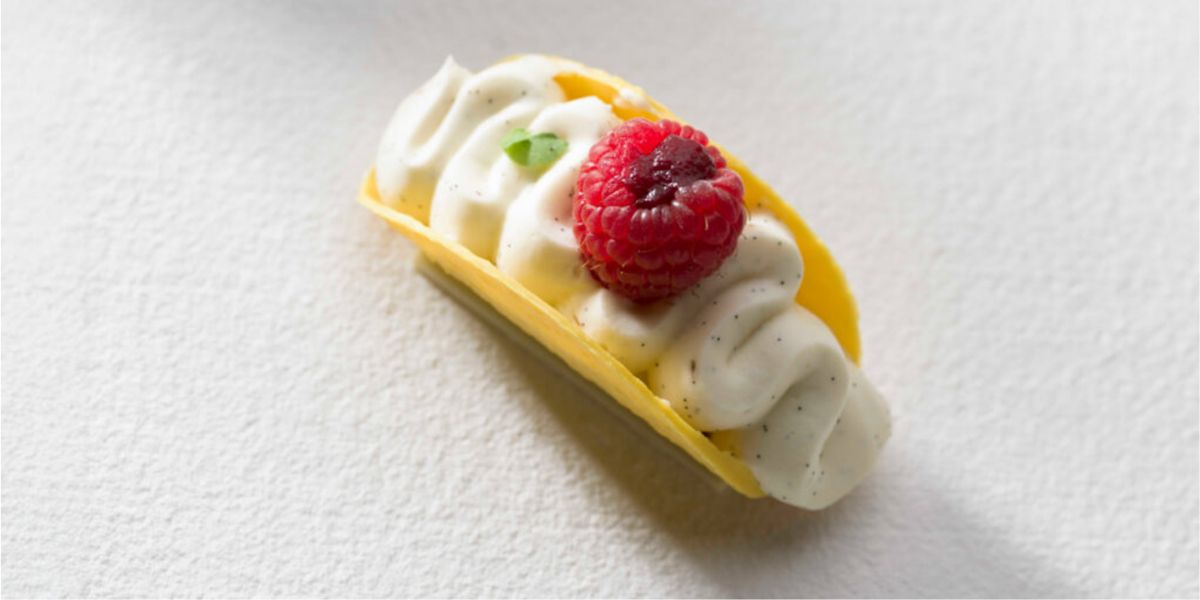 A taco-shaped white chocolate shell filled with piped cream and topped with a raspberry